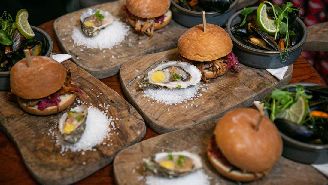 Four burgers on lush buns with a wooden stick placed in the middle to keep them standing upright all on different bread boards with an oyster on the side as well as a side salad.