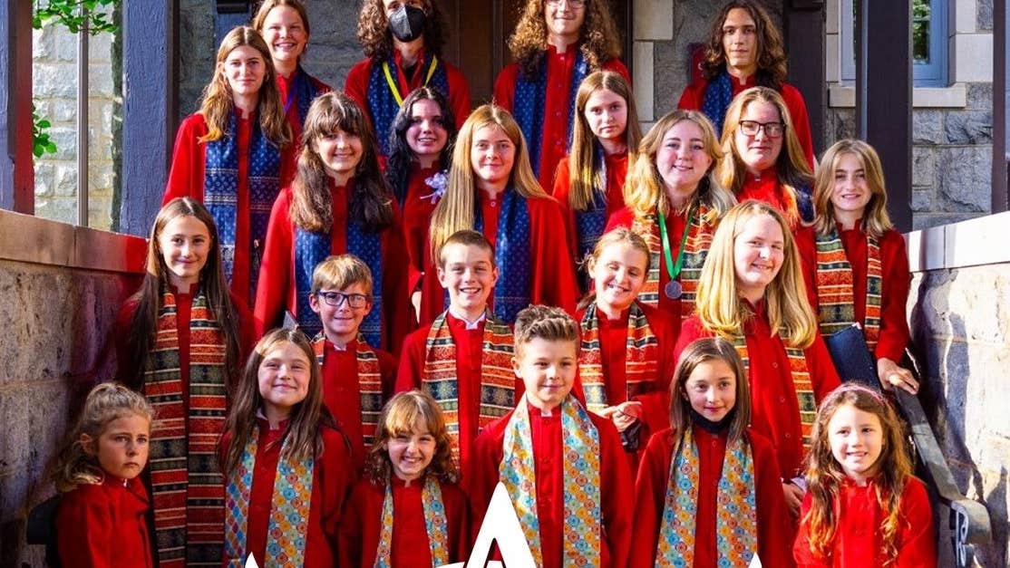 Group of children tiered, standing on steps, dressed in red gowns with multicoloured scarfs, smiling at the camera.