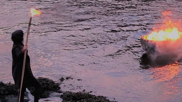 A person in a cloak holds a lit torch, watching a pire blaze in the middle of a misty river.