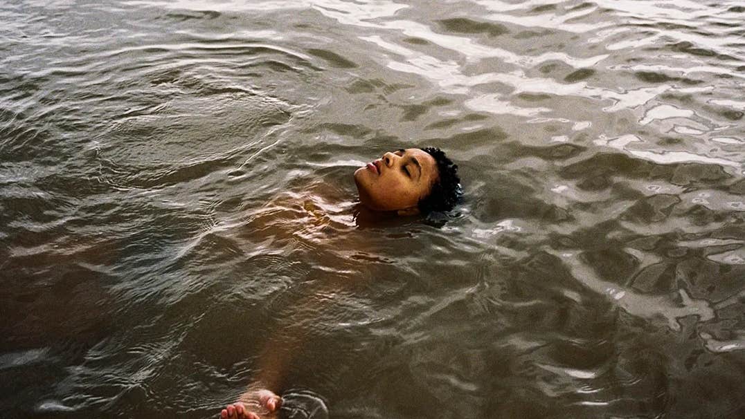 A young person is floating on their back in grey water with head and one hand visible above the water.