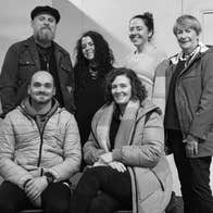 A group of smiling people with 2 seated, the rested standing, some wearing coats indoors.