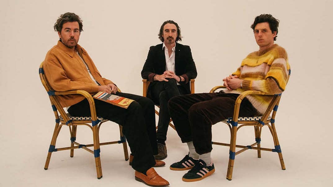 3 men seated in huddle, looking serious, against plain, beige background.