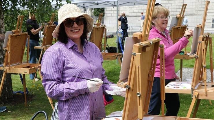 Easels set up outdoors with smiling woman in a hat painting