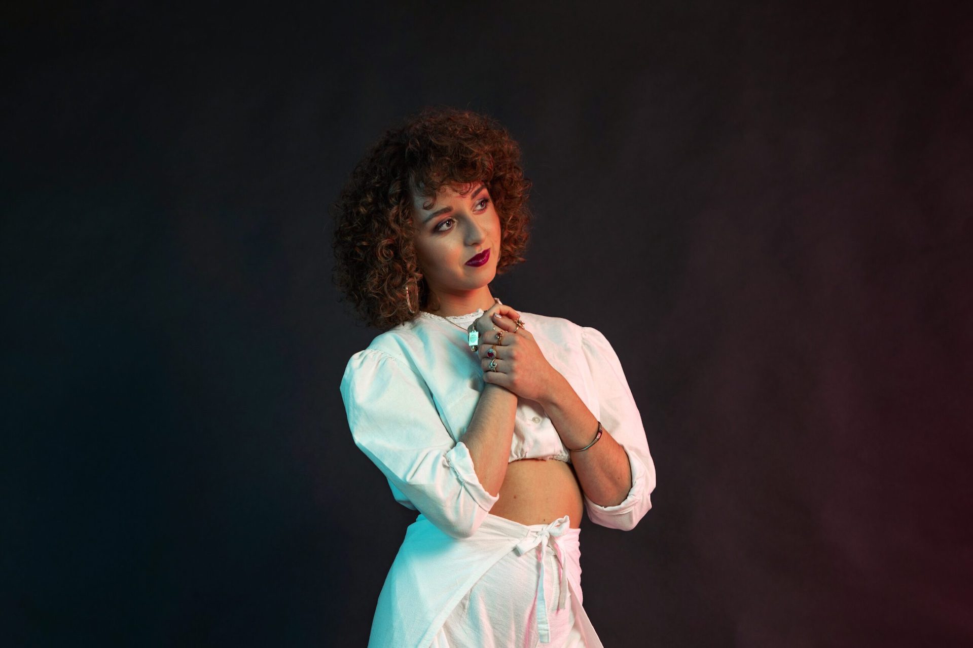 A woman dressed in white is clasping her hand together up in front of her chest, looking away to her left looking thoughtful against black background.