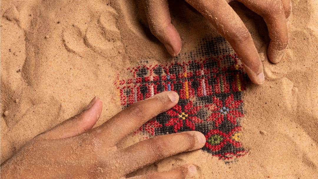 Looking down at a pair of hands digging out a small square of red and black patterned cloth from sand.