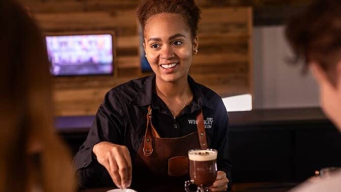 A smiling person in apron with 2 drinks in front on a wooden counter.