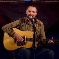 A man seated playing a guitar, singing into a mic on a stand