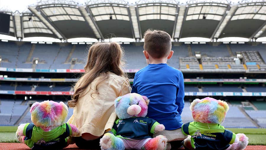 Rear view of 2 children seated on the ground looking at stands in a large stadium with 3 colourful bears behind them.