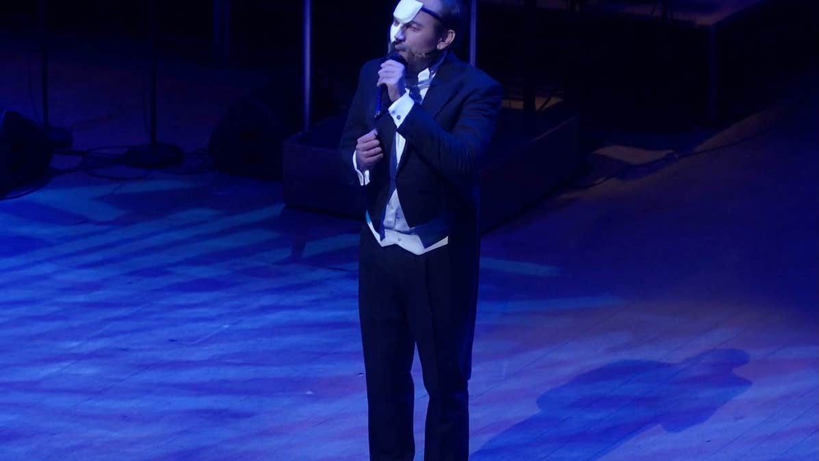 A man in evening dress singing into a mic on a dark stage, half of his face covered with a small mask.