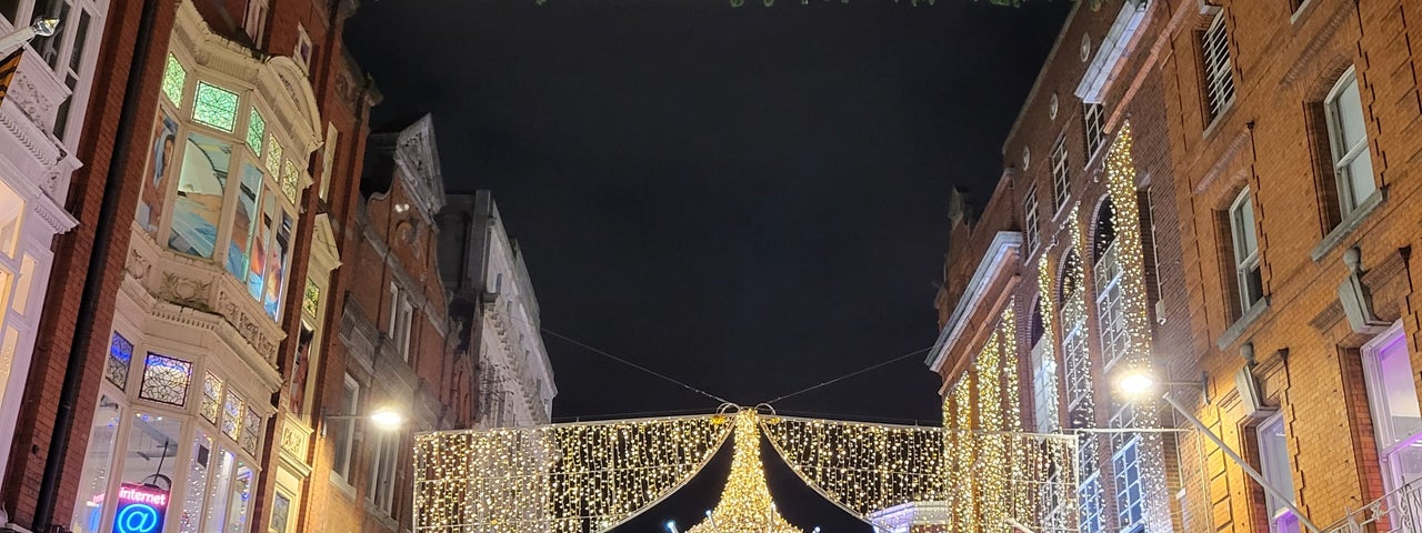Looking down a shopping street with buildings in a street at night with yellow christmas lights across the between them.