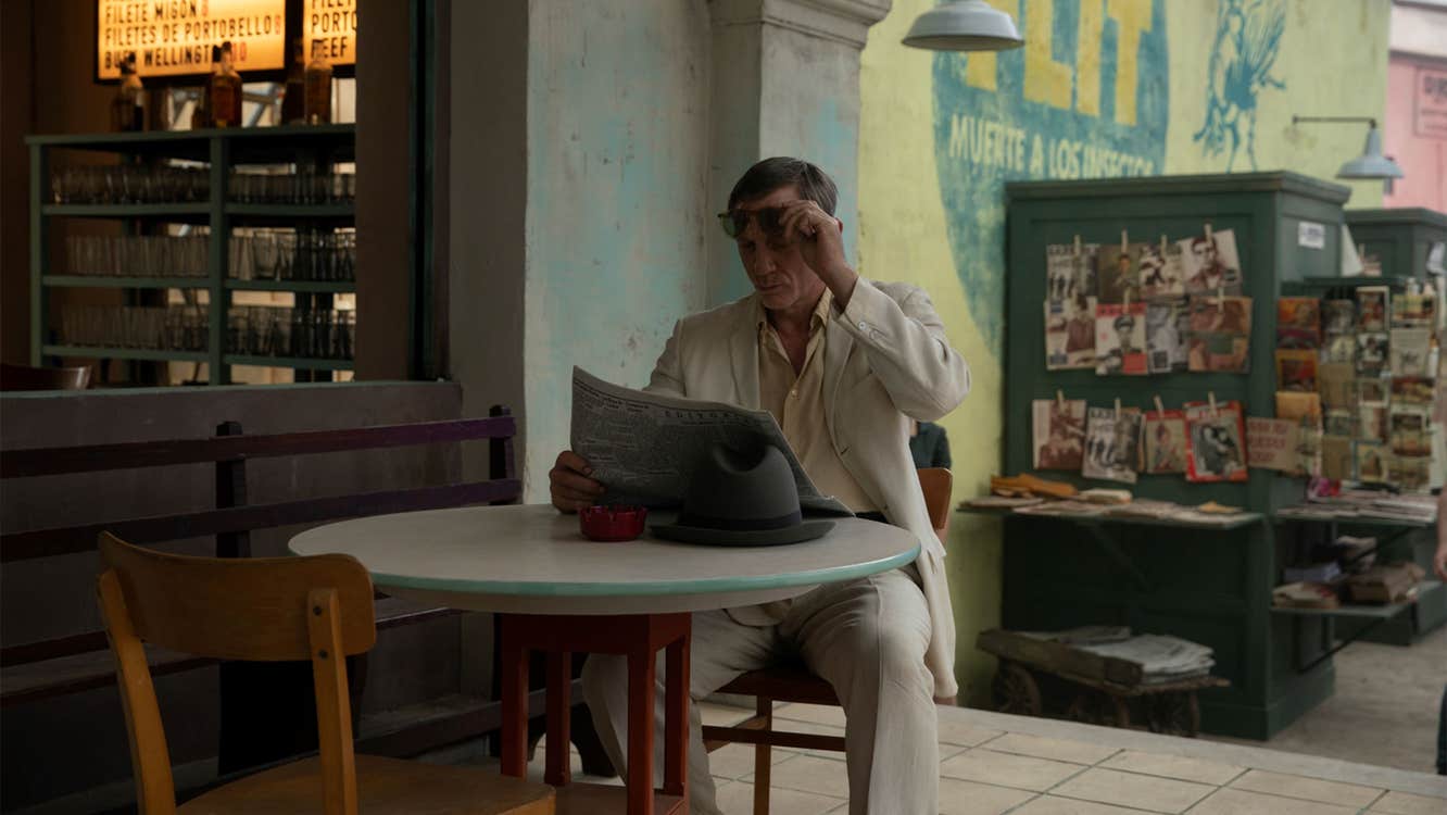A late middle-aged man in a white suit is reading a newspaper at a cafe table.