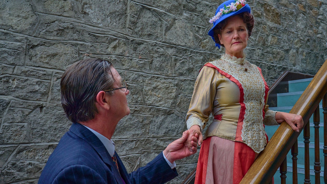 A woman in early 1900 century dress and hat is on staircase, holding out hand to man at the bottom of the stairs.