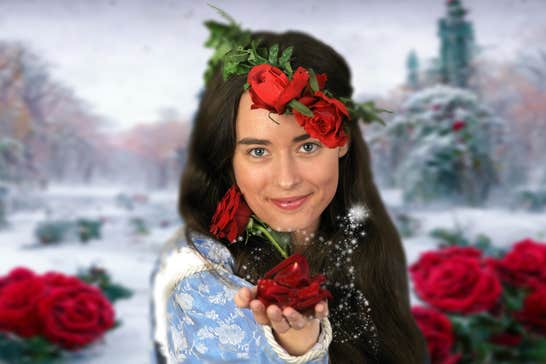 A woman with dark hair is smiling, holding forward a dark red rose in her outstretched hand with dark red roses and green leaves behind her against a snowy outdoors scene.