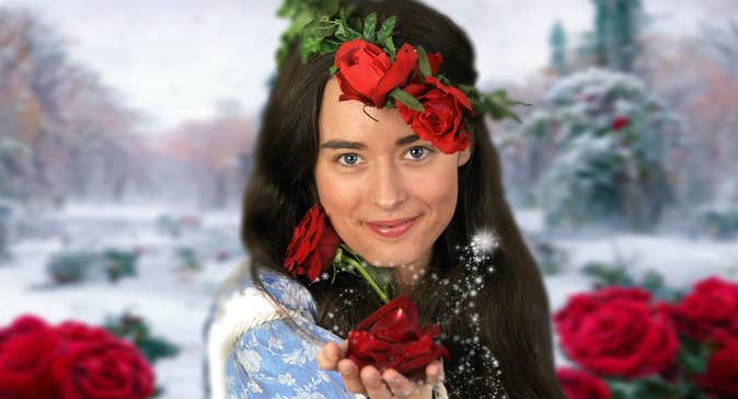 A woman with dark hair is smiling, holding forward a dark red rose in her outstretched hand with dark red roses and green leaves behind her against a snowy outdoors scene.