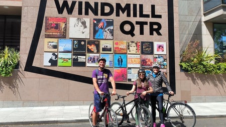Three people on bikes outside a building with album covers displayed on it