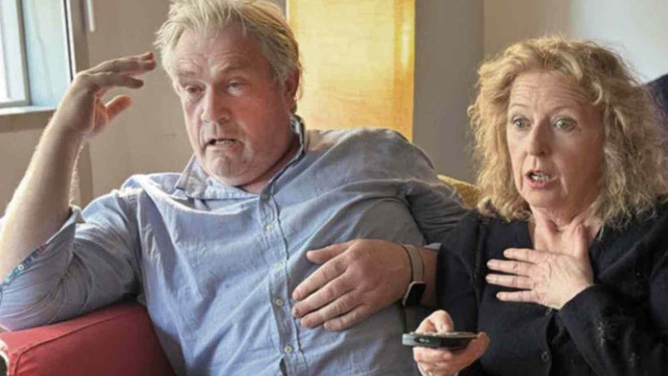 Man and woman sat close together on a sofa, both looking ahead with shocked expression, the woman is holding up a remote control.