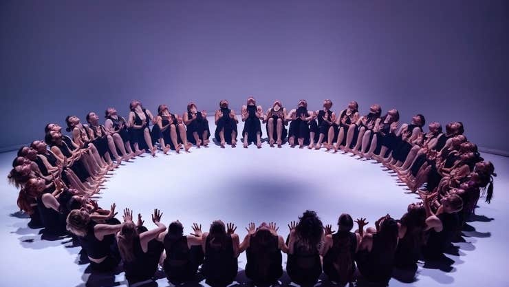 A large group of dancers seated on a white floor in a large circle.