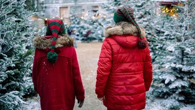 Rear view of 2 children in red coats walking with snow covered christmas trees