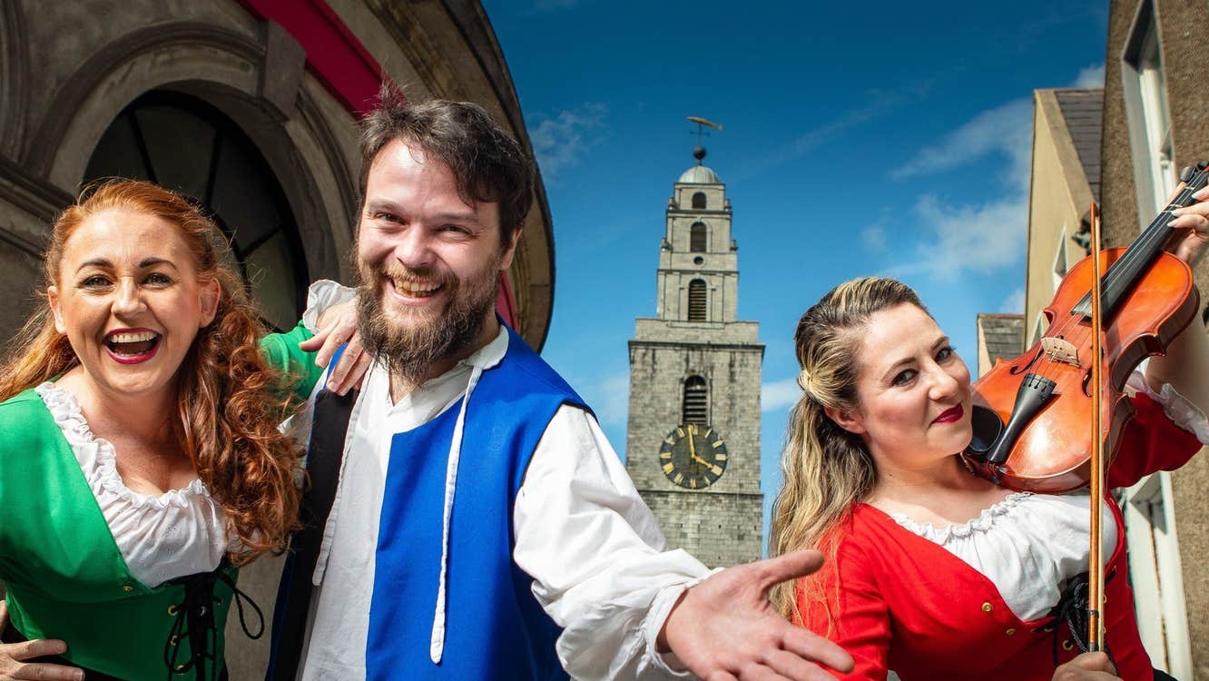 Smiling actors with one playing a fiddle and a clock tower in the background