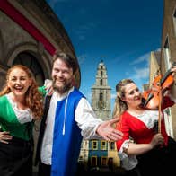 Smiling actors with one playing a fiddle and a clock tower in the background