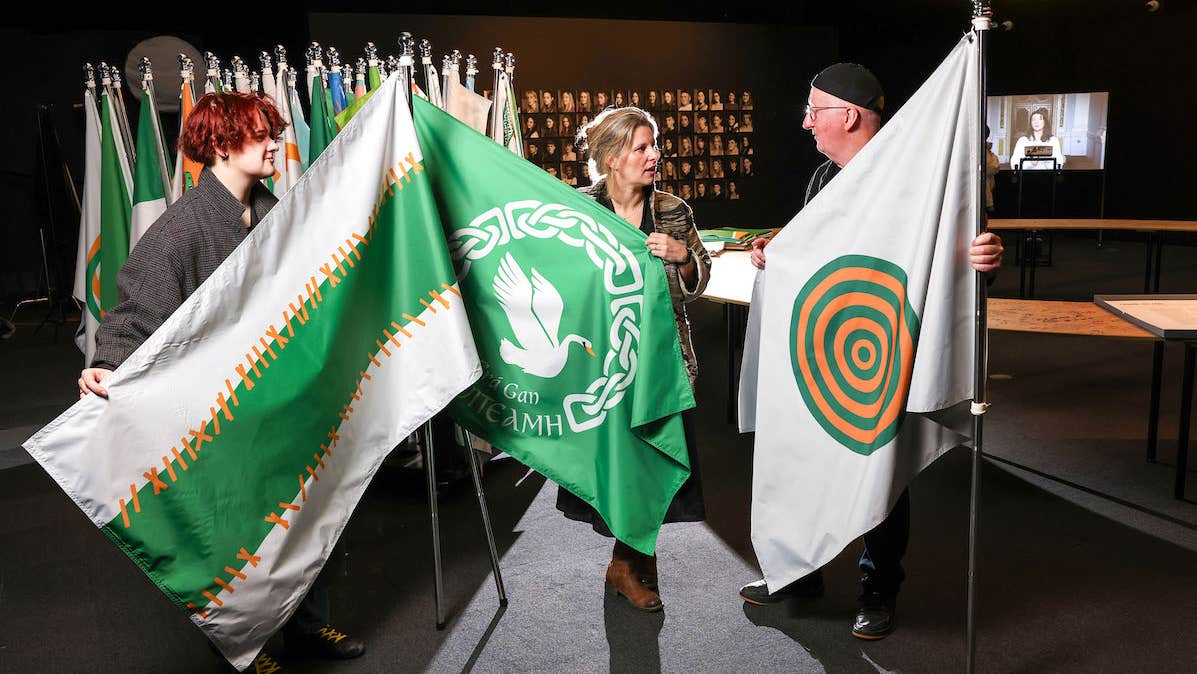 3 people in a discussion, each holding a large flag on a pole with a large number of flags behind