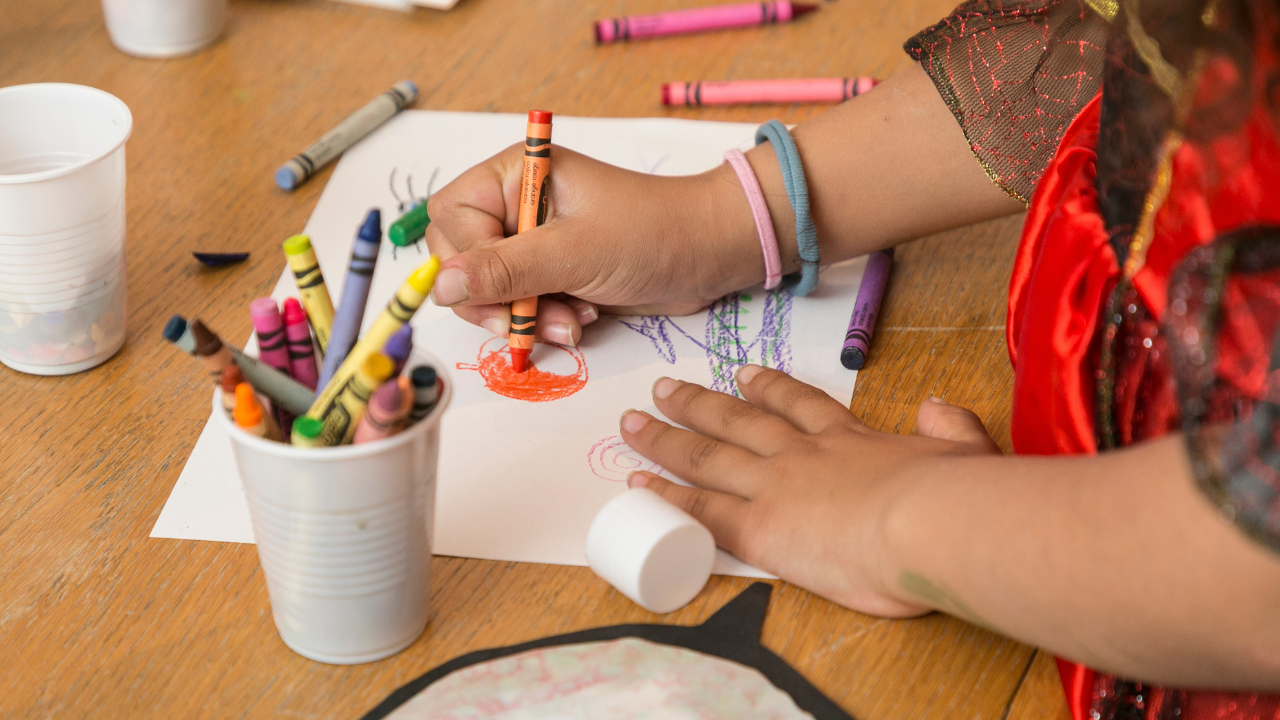 On a table, a child's hands and arms are visible, drawing on paper with crayons.