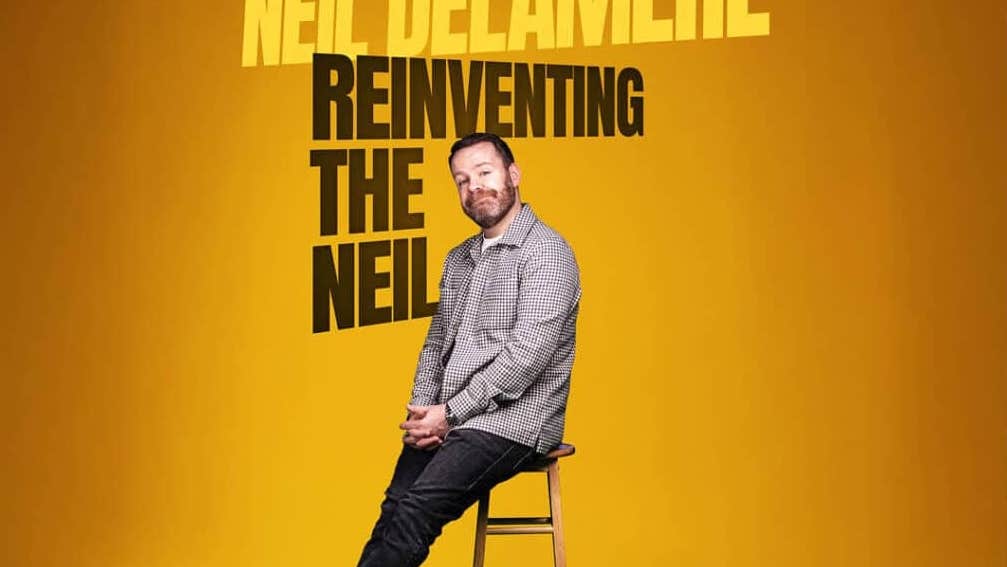 Award-winning Irish comedian Neil Delamere, a smiling man perched on the edge of a tall stool.