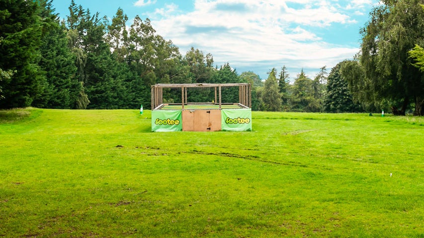 A green field area surrounded by trees with a wooden structure in the distance