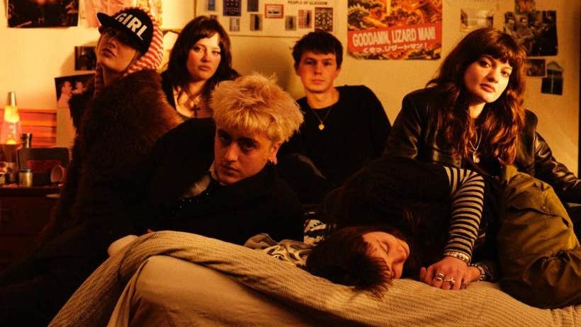 A group of 6 people seated on a bed in a room with poster on the wall behind