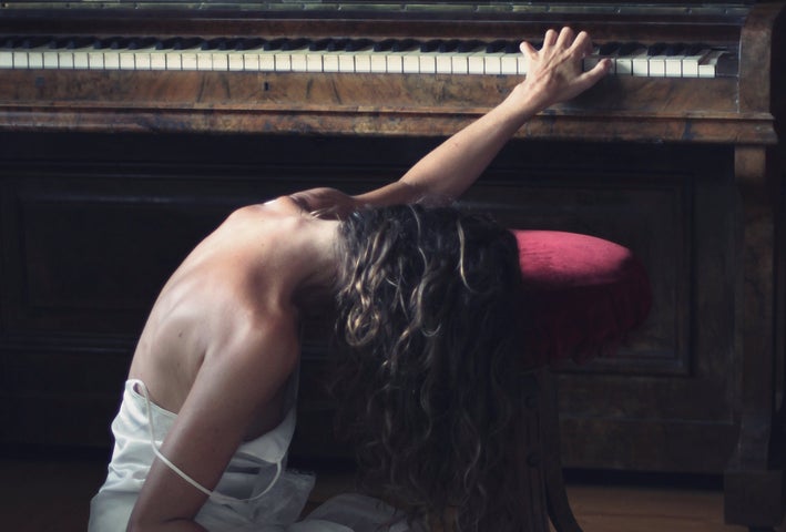 A person is sitting on the floor with one hand reaching up to the keyboard of a piano beside them, their face is covered by hair.