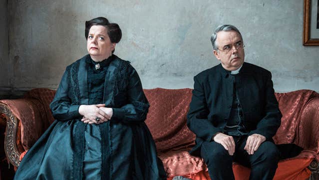 A serious looking man and woman dressed in black seated on a large sofa