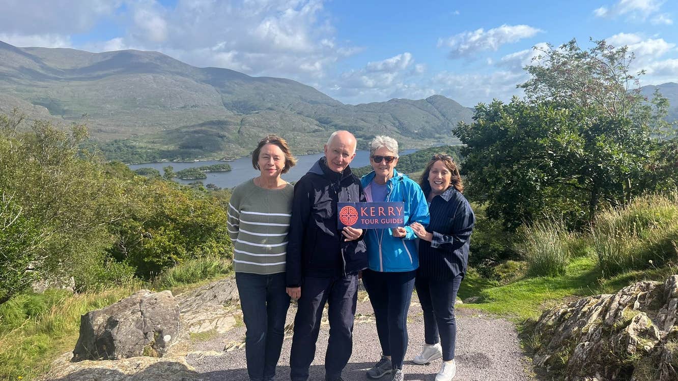 Four people on a path holding a sign with lakes and mountains in the background