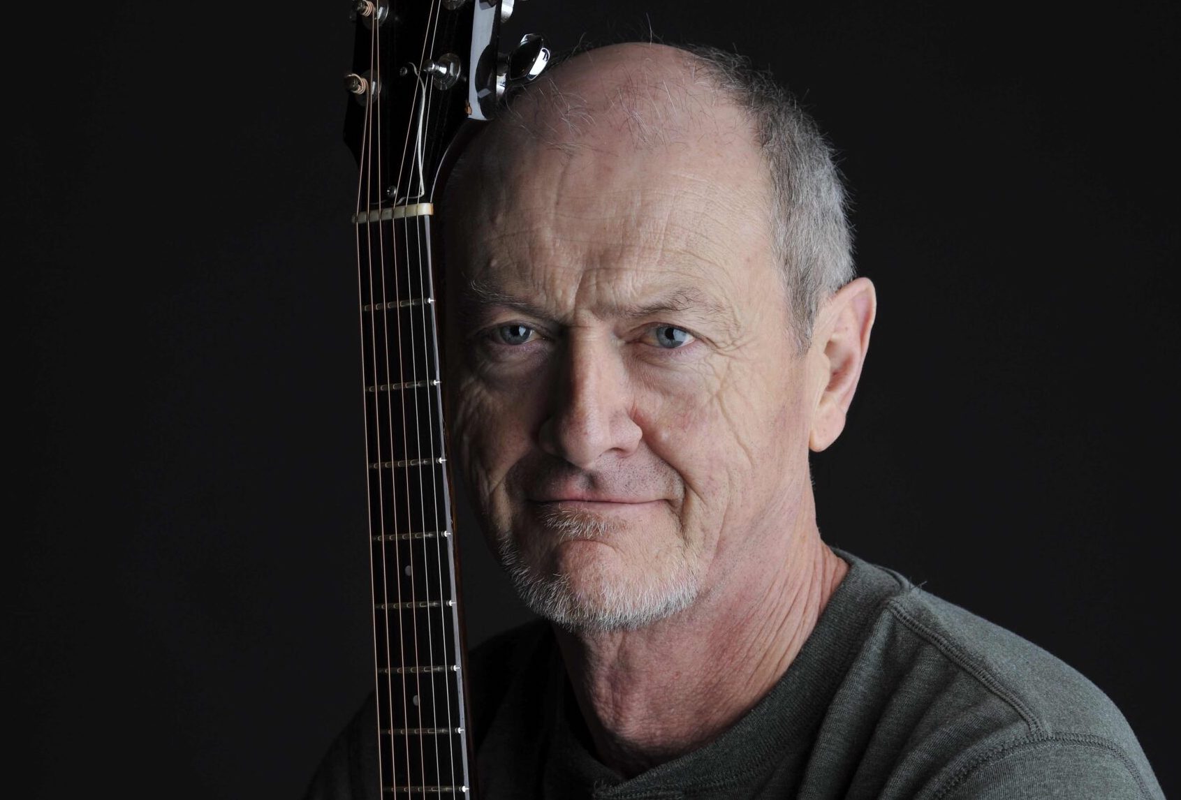 Headshot of man holding a guitar upright beside him so only the neck is visible beside his face, against black background