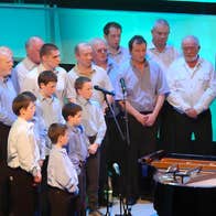 A large group of men and boys all wearing pale, plain coloured shirts, gathered around a man seated at a grand piano.