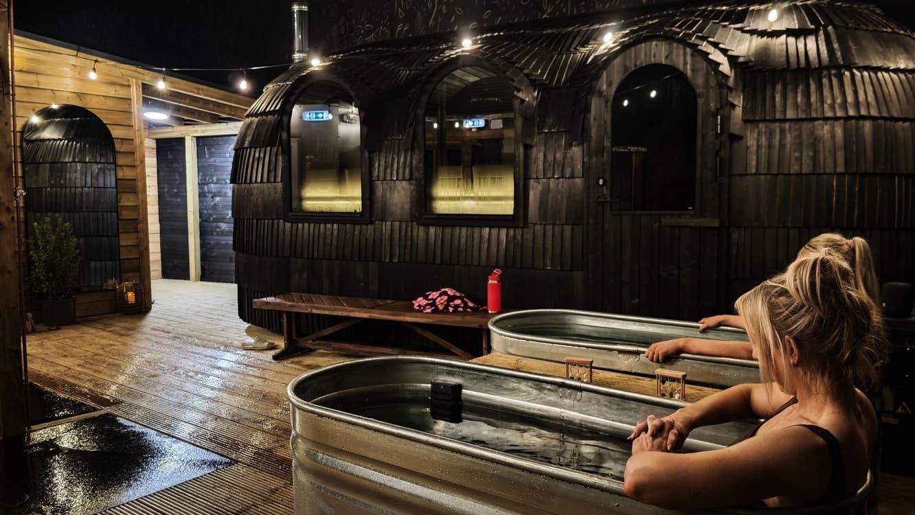 Two people sitting in cold plunge baths with mobile saunas to the right