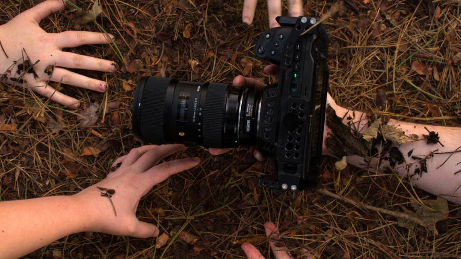Looking down at 5 hands reaching for a camera with large lens in brown earth and twigs.