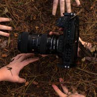 Looking down at 5 hands reaching for a camera with large lens in brown earth and twigs.