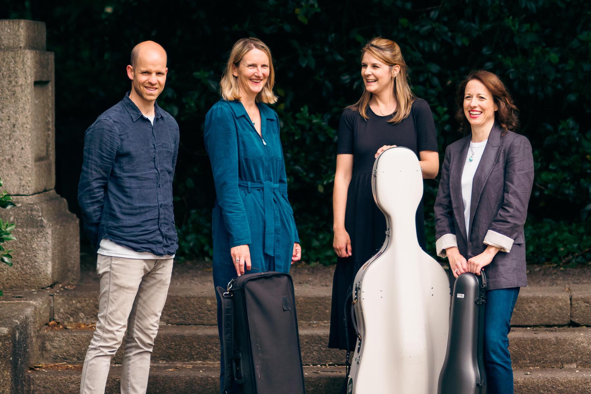 Line of 4 smiling people, some holding musical instrument cases outdoors with stone pillar behind.