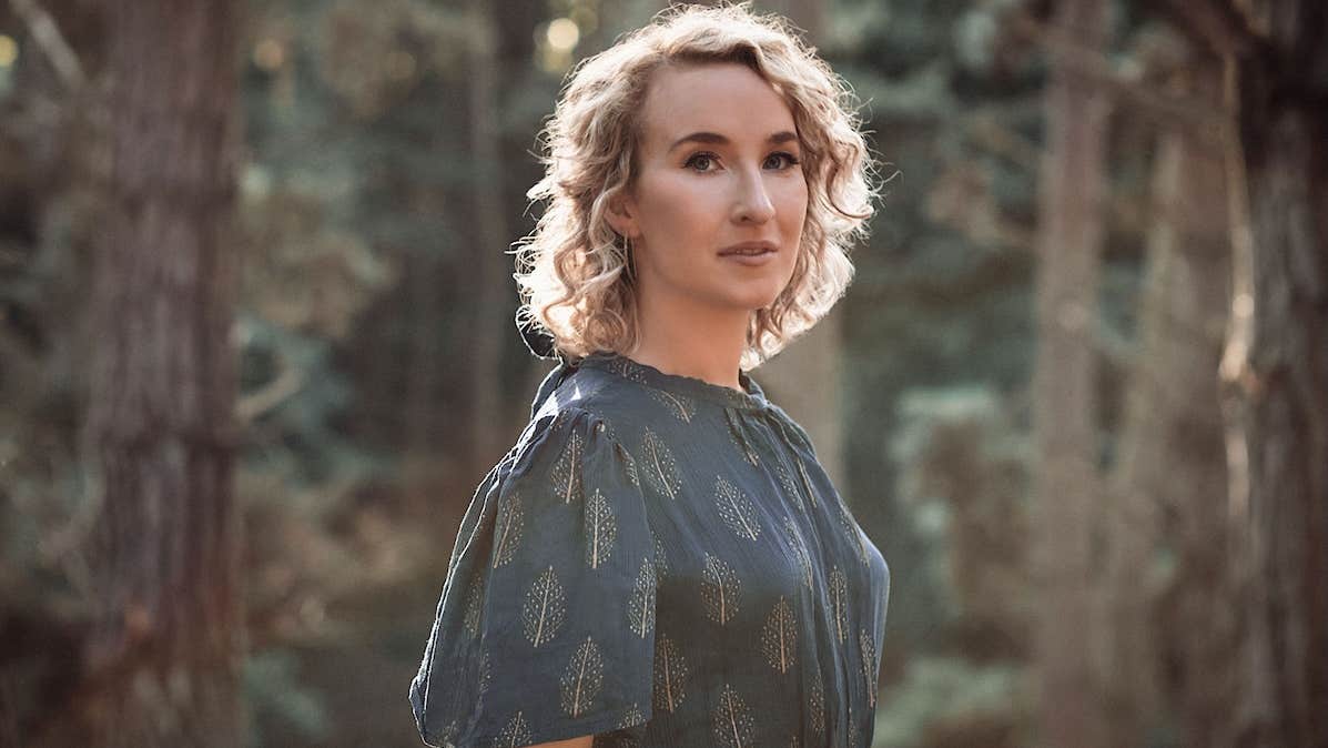 Slightly side profile on woman in dark, patterned dress with blurry trees in background.