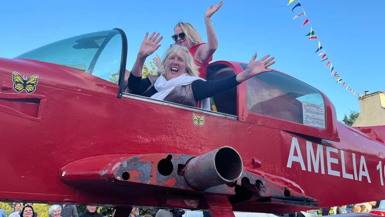 2 smiling and waving women in the cockpit of an old red small aeroplane