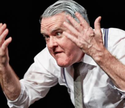 Mikel Murfi stars in The Man In The Womans Shoes. A man in shirt and tie has both hands raised, looking aggrevated.