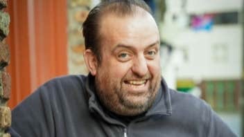 A smiling man in grey fleece is leaning on a window ledge outdoors.