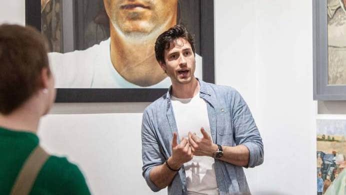 A man is talking and gesturing with his hands, standing in front of pictures on a wall.