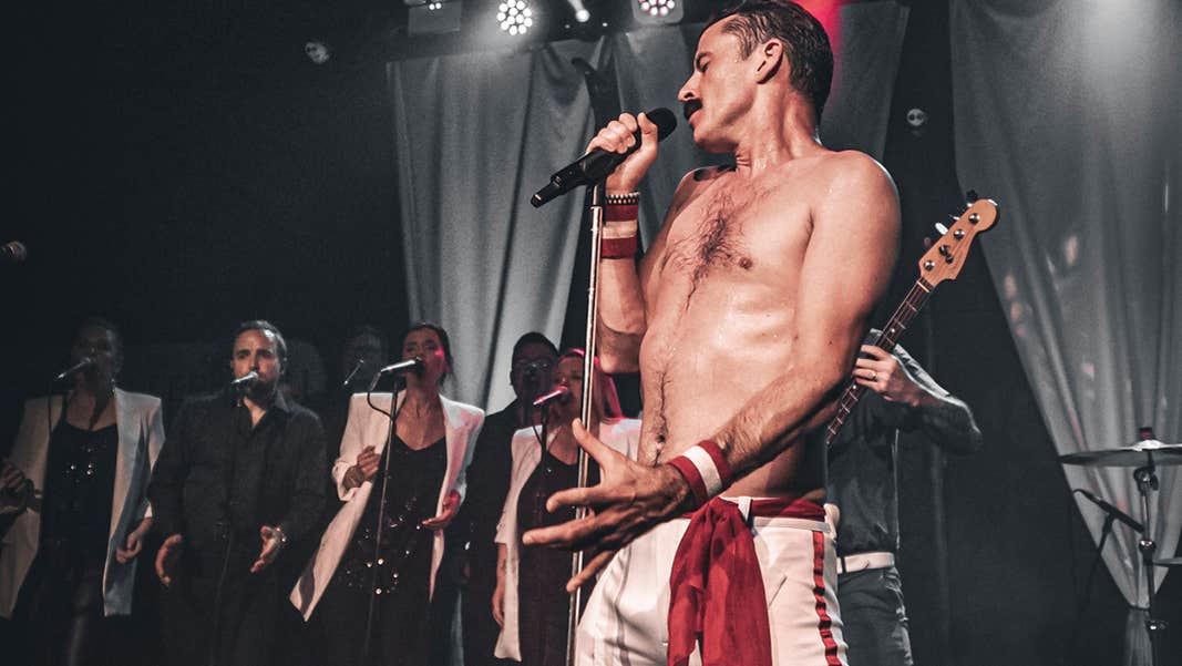 A shirtless man on a stage is singing into a mic with line of singers in the background.