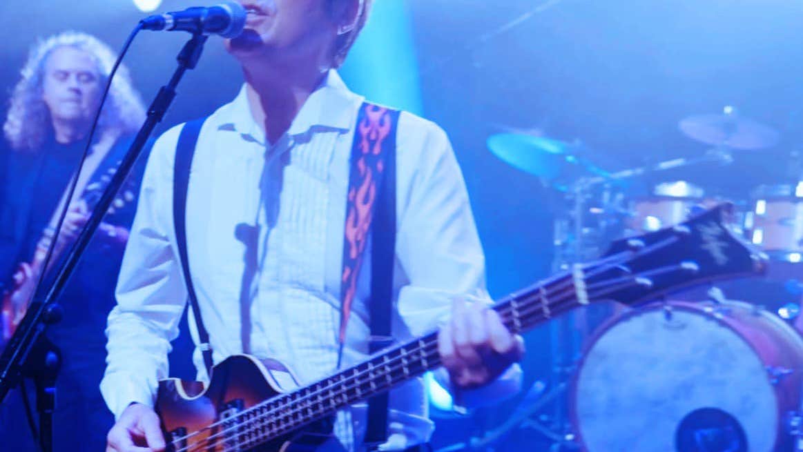 A man playing a guitar singing into a mic on a stand with band members behind