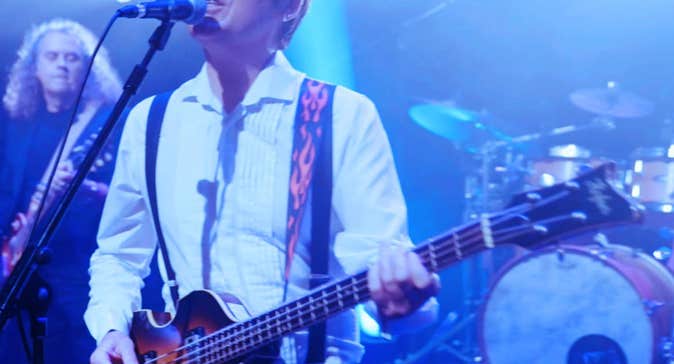 A man playing a guitar singing into a mic on a stand with band members behind
