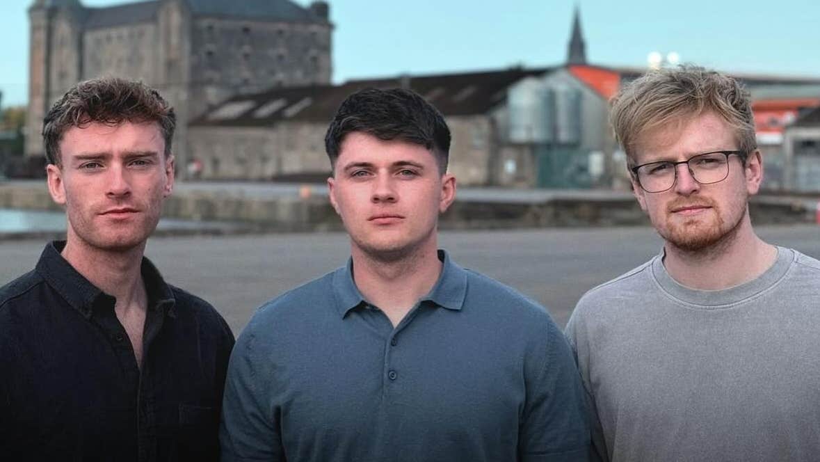 3 men standing close together with water and buildings in the background.