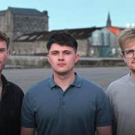 3 men standing close together with water and buildings in the background.