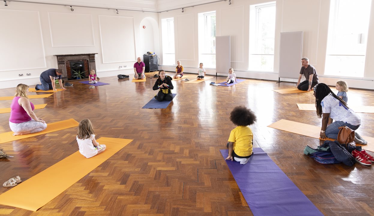 Family yoga at the National Museum of Ireland- Decorative Arts & History. A large, plain room with wooden floor, adults and children are sitting on yoga mats on the floor, spread out around the room.