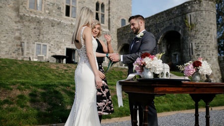 A bride and groom standing at a table with flowers outside a period house